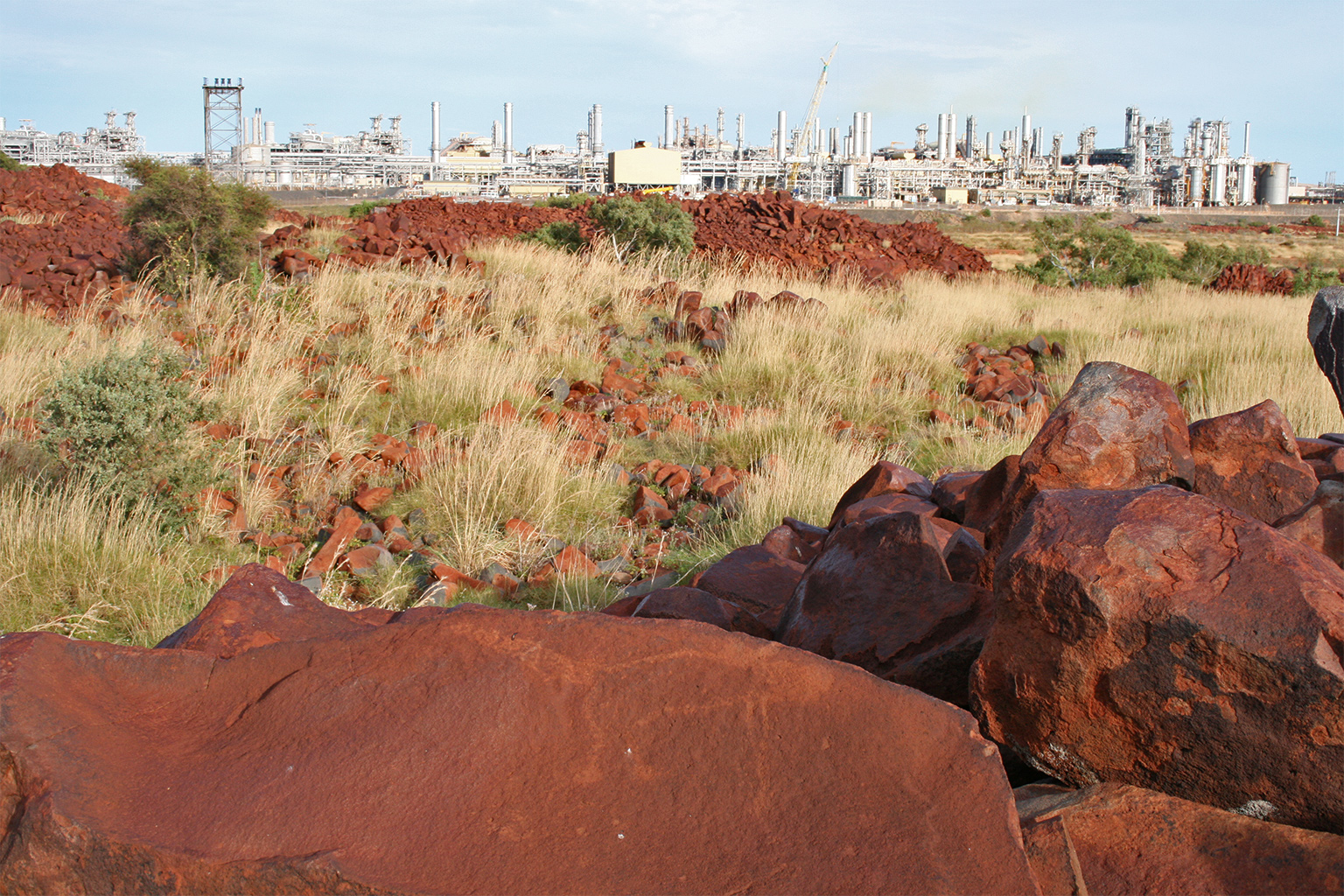 Woodside Energy factory can be seen beyond the red rocks of Murujuga. The petroglyph of a kangaroo can be seen on the rock.